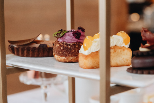 A tiered tray displaying various pastries, including cakes with cream and fruit toppings, and scones with raisins. The setting is elegant and inviting.